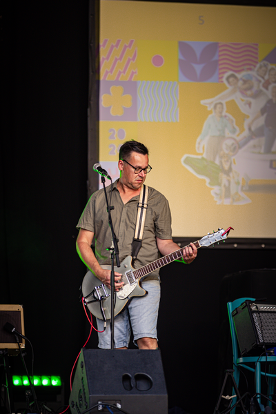 A man in a brown shirt and sunglasses is playing a silver guitar on stage with a yellow background featuring children.