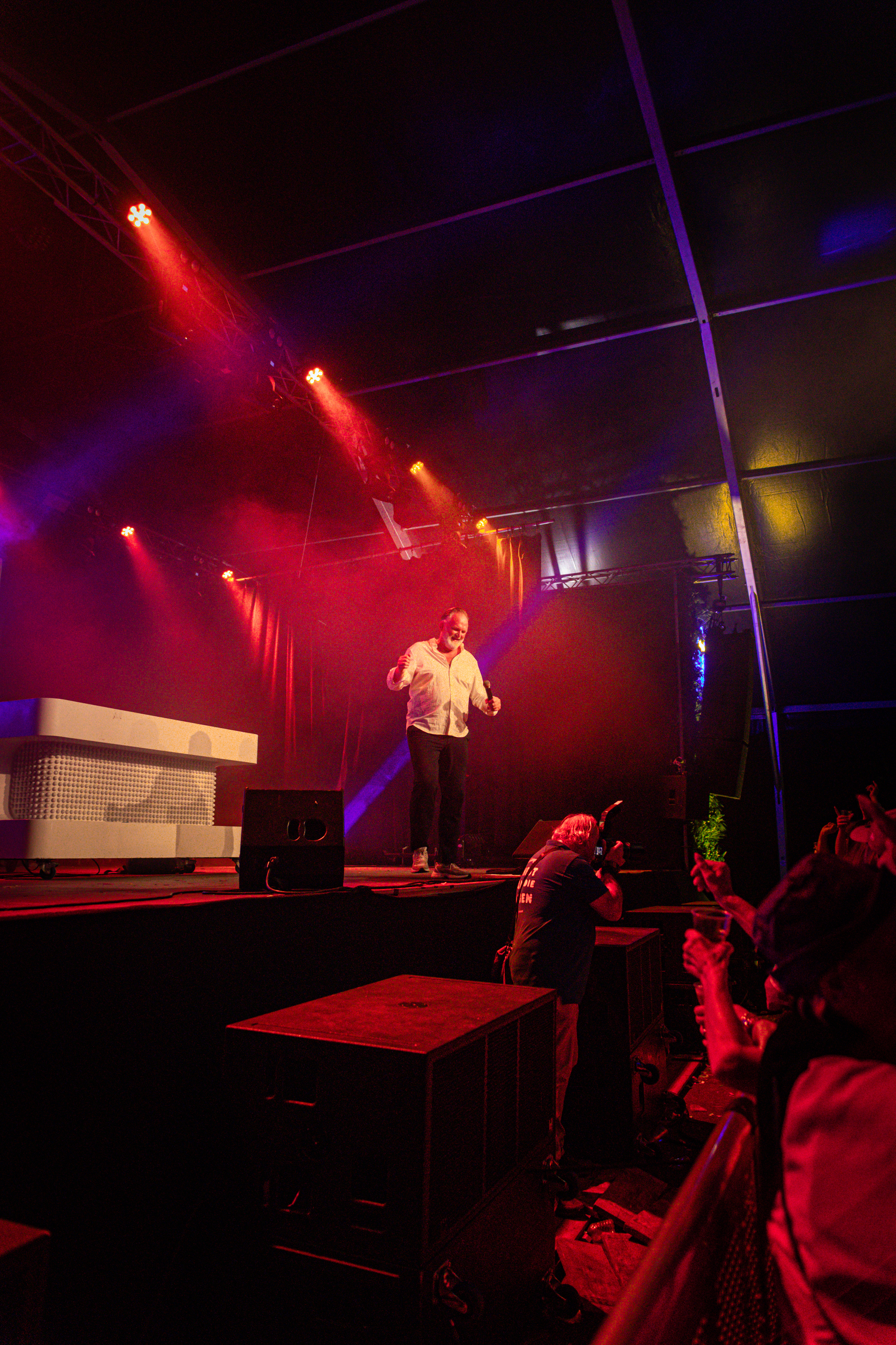 A man is standing on a stage with red and white lights, wearing a white shirt.